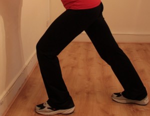 Standing leaning on wall with one leg lengthened behind for calf stretchh
